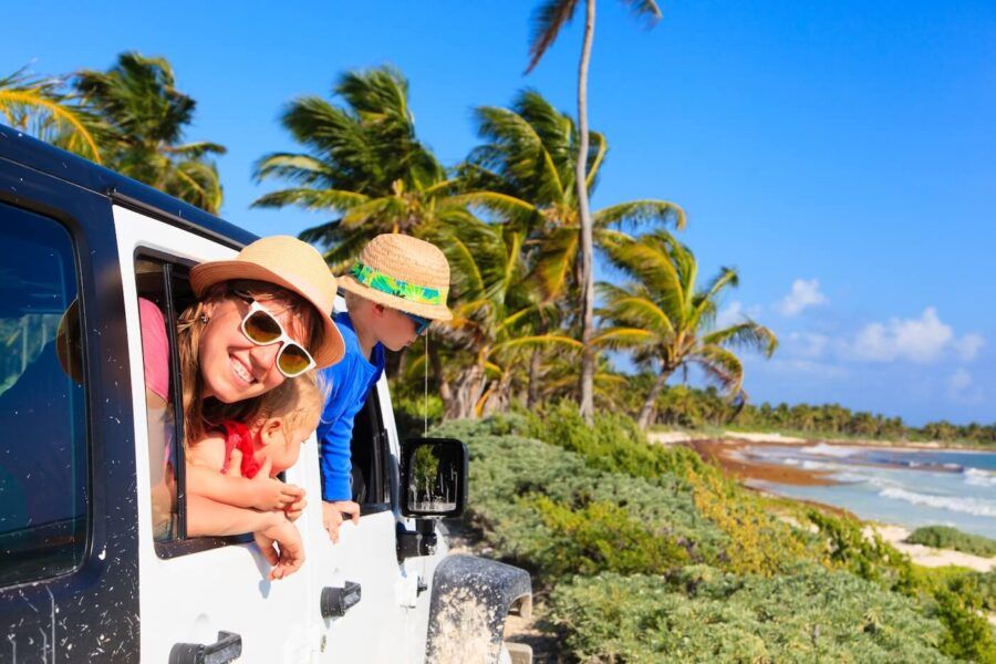 A happy mother with two toddlers in sunglasses and straw hats sticking out of the car window near a beautiful tropical beach