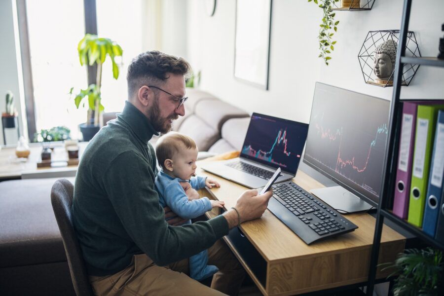 Man looking at currency trading app on his smart phone from his home office while holding a baby.