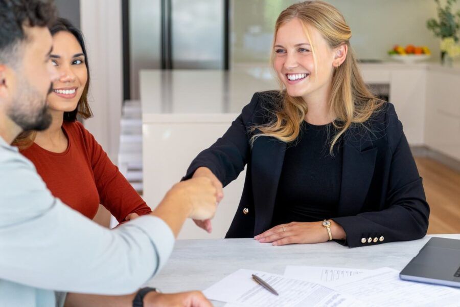 Smiling female mortgage broker shaking hands with a male client, a smiling young woman sitting to him