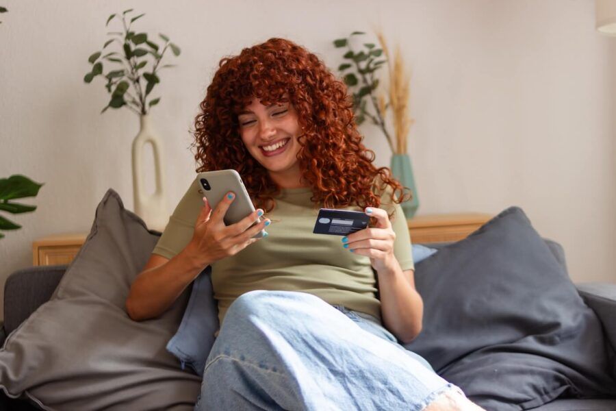 Happy woman is enjoying online shopping from the comfort of her couch, using her smartphone and credit card to make purchases