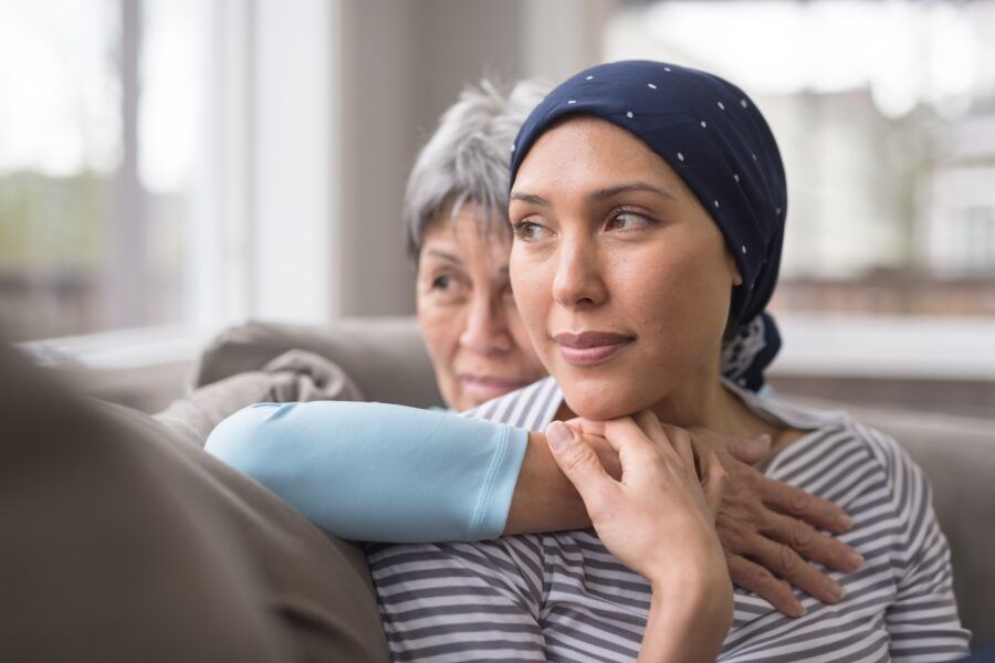 A woman wearing a headscarf and fighting cancer sits on the couch with her mother. She is in the foreground and her mom is behind her, with her arm wrapped around in an embrace, and they're both looking out the window.