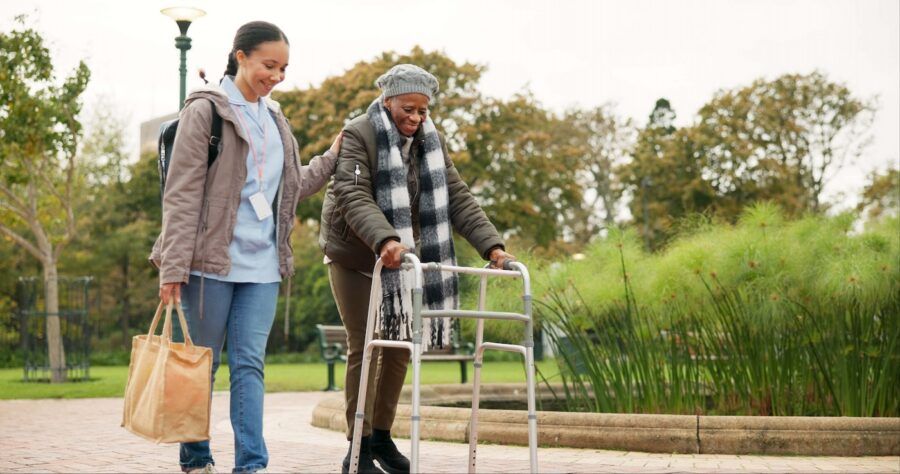 A long term care nurse walking with a patient using a walker in a nice park.