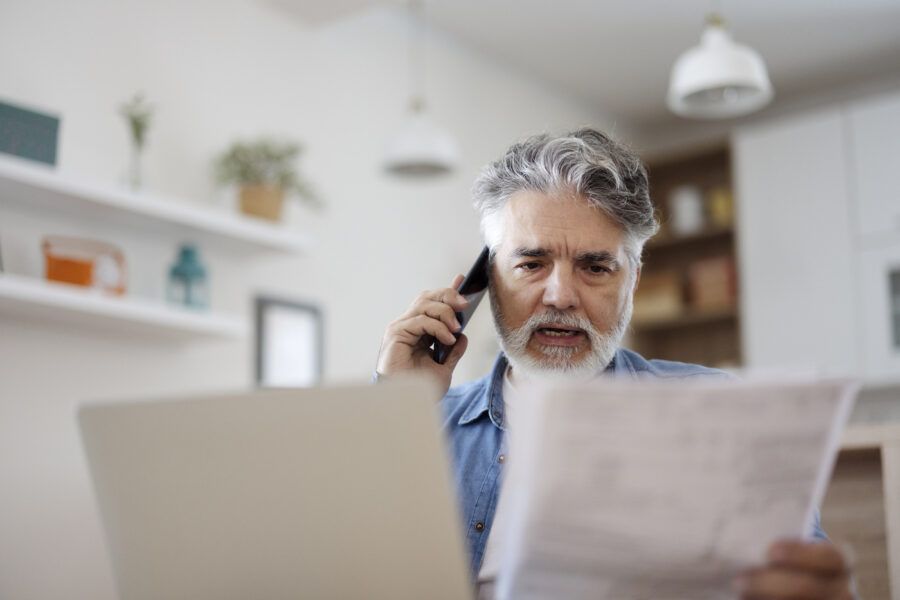 Stressed mature man holding paper and talking on phone at home.