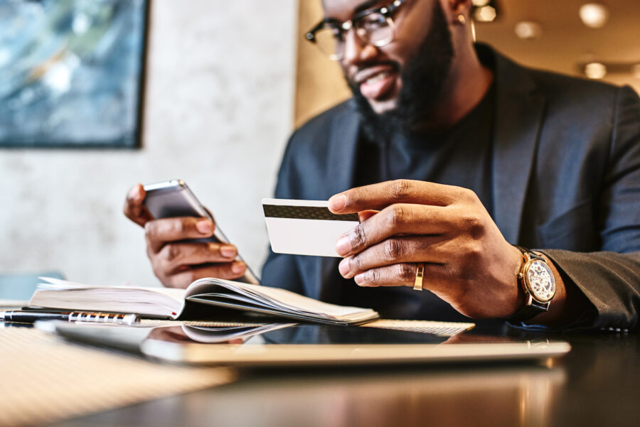 Smiling man holding his credit card and checking its benefits on his mobile phone while sitting at a desk full of papers