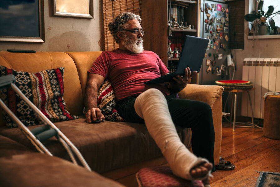 Mature man with broken leg in plaster cast on sofa checking the status of his SSDI