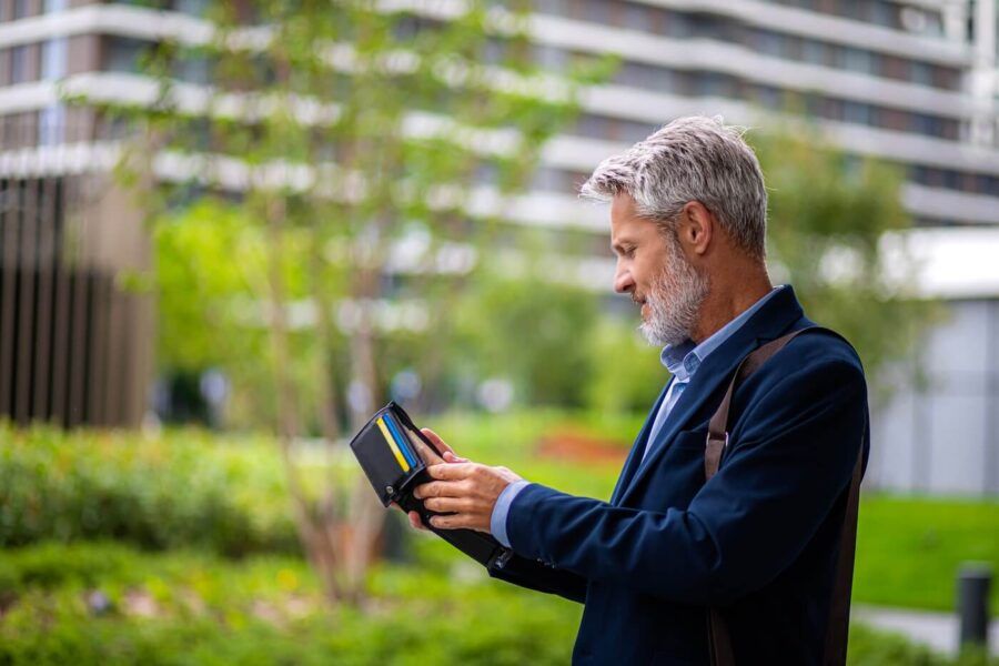 Mature man in business attire standing outdoors near modern office buildings, holding an open wallet with visible payment cards and reviewing its contents.
