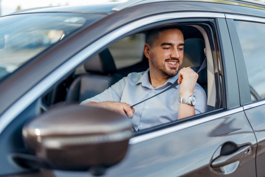Smiling man seated in the driver's seat of a vehicle fastening a seat belt before driving. The image shows the interior of a modern car, including the side mirror, door frame, and front cabin area.