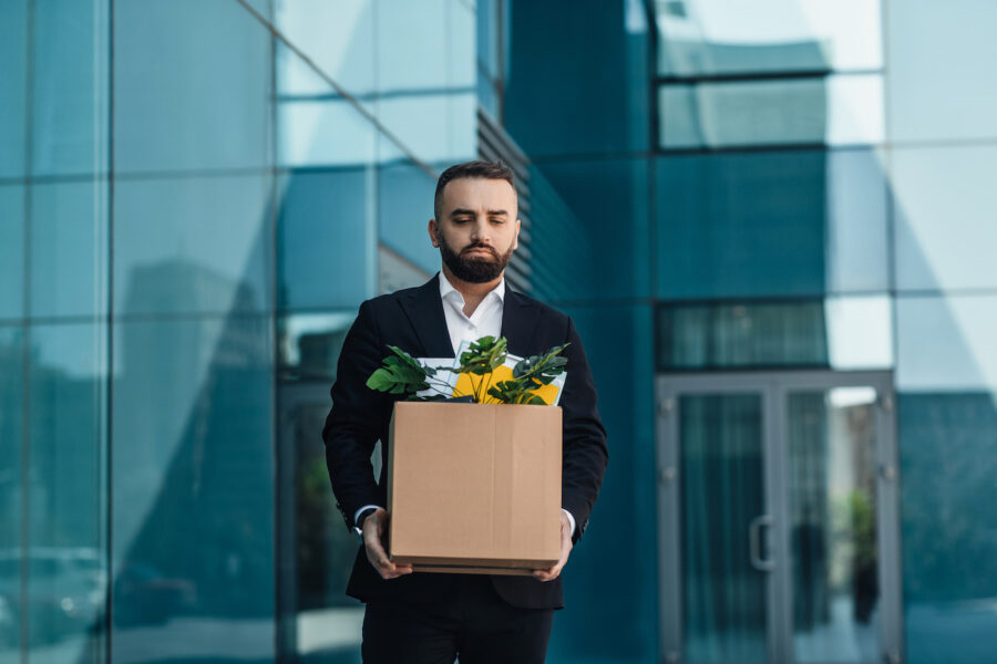 Upset male office worker holding box with things and leaving office building