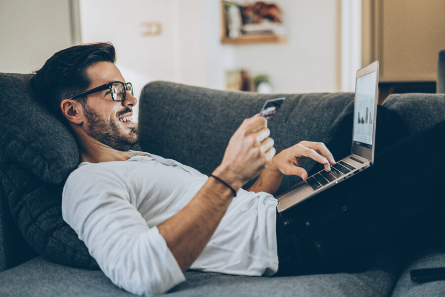 Smiling man lying on the couch and shopping online with credit card and laptop