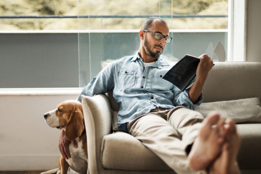 Bearded man comfortably sitting on a coach reading a book and petting his dog, living alone.