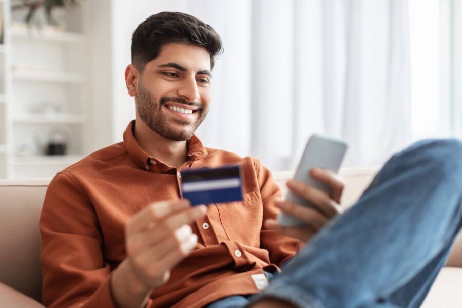 Smiling young man relaxing on a sofa at home while holding a credit card and using a smartphone, illustrating online shopping, mobile banking, or digital payments in a bright living room setting.