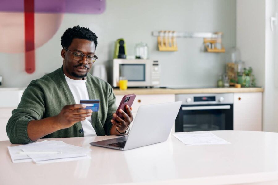 Focused man seated at a kitchen table holding a credit card and using a smartphone beside an open laptop. Documents are spread across the table, with kitchen appliances, countertops, and cabinets visible in the background.