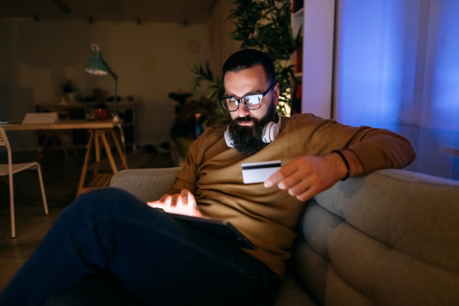 Man holding a credit card sits on his couch as he shops online. His laptop screen glares on his glasses as he sits in the dark.