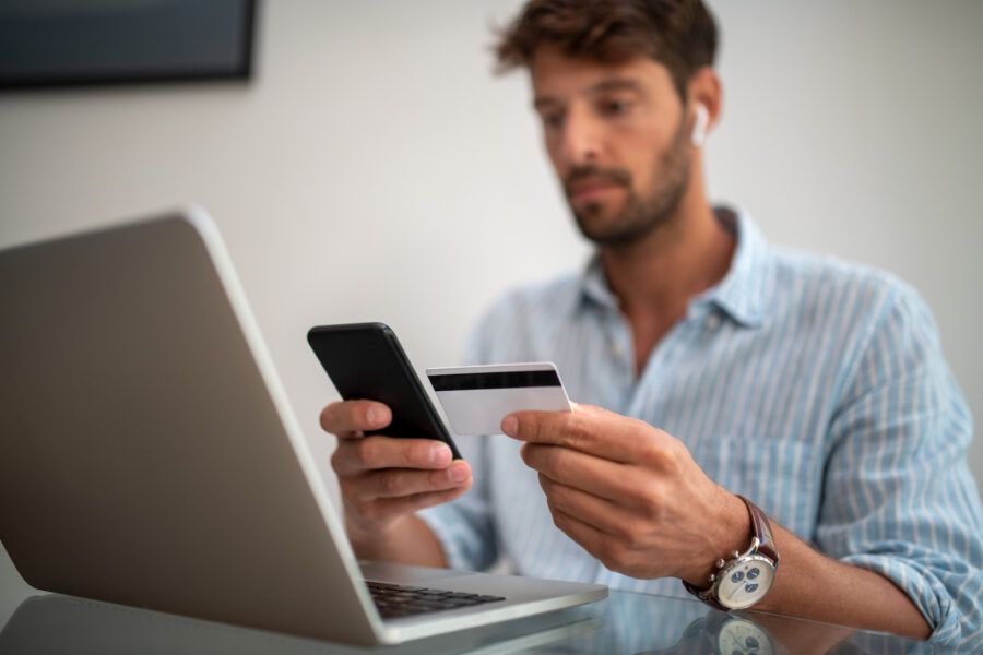 Close up of a man paying a credit car bill online, holding his phone and credit card in front of a laptop.