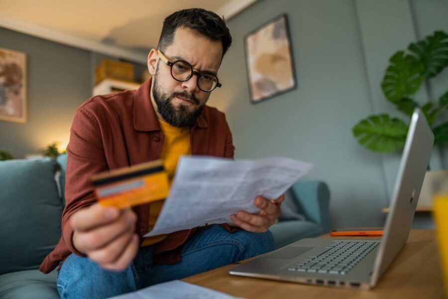 Concerned man seated in a living room reviewing a printed bill while holding a credit card, with a laptop open on a coffee table nearby. The home setting includes a sofa, houseplants, framed wall art, and soft indoor lighting.
