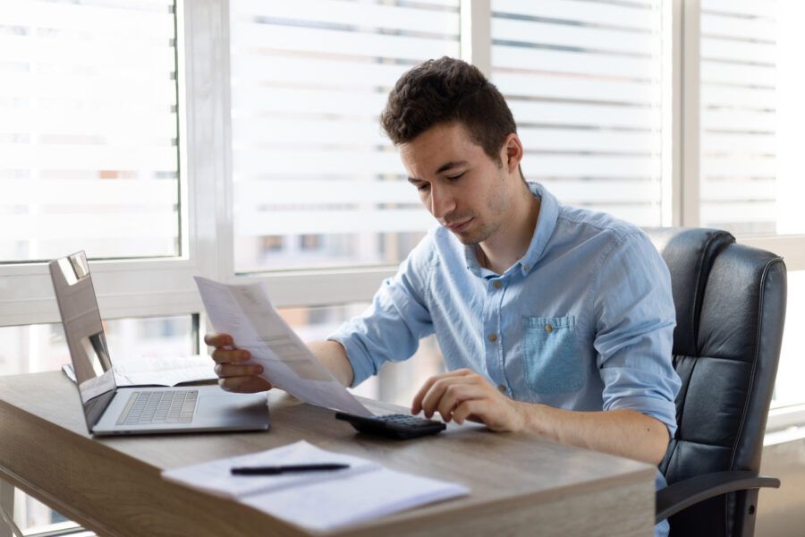 Young man reviewing debt consolidation loan, sitting at a desk and holding papers in front of a laptop.