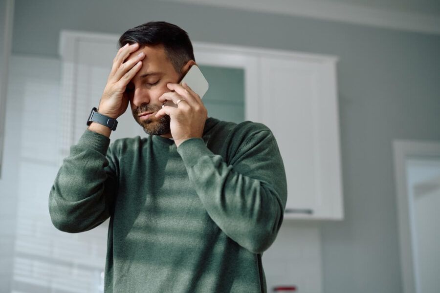 Upset man standing in a home kitchen holding a smartphone to one ear, with one hand resting on the forehead, wearing a green sweater and a smartwatch, with cabinets and window blinds visible in the background.