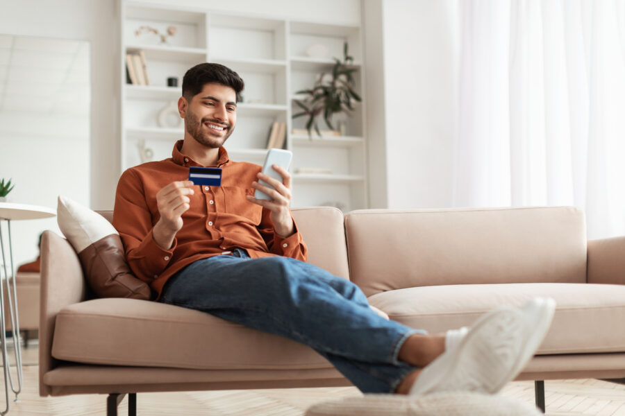 Man sitting on the couhc and smilingholding a credit card, learning more about credit by reading on his phone