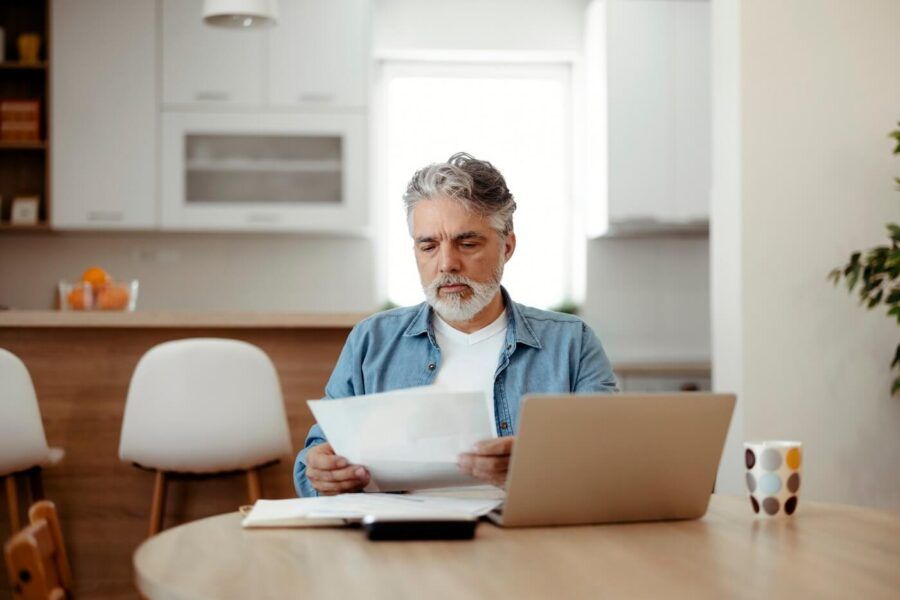 Focused mature man seated at a dining table reviewing printed documents beside an open laptop, with a notebook, smartphone, and coffee mug on the table in a bright home kitchen setting.