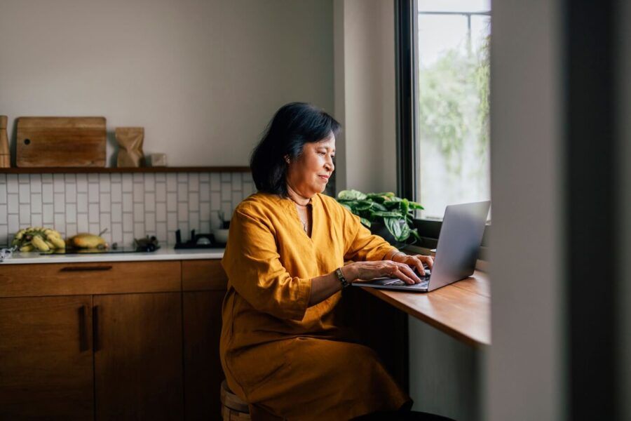 Mature woman seated at a kitchen counter beside a window, using a laptop computer in a bright home setting. Wood cabinetry, open shelving, a tile backsplash, houseplants, and natural light are visible in the background