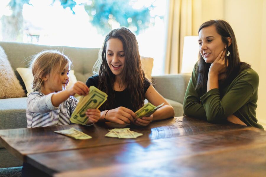 Young mom teaching her two elementary-age daughters about money, daughters counting bills on the coffee table.
