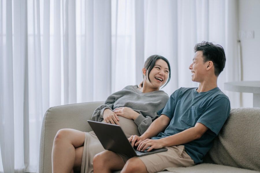 Happy couple sitting on a couch, the man is using his laptop while smiling at the pregnant woman