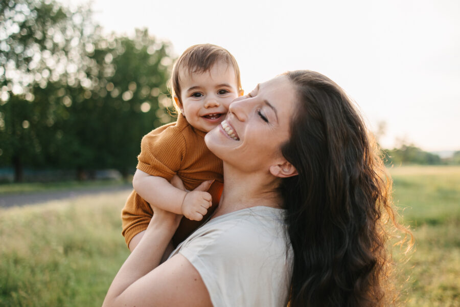 A close-up of a young mom holding her baby boy in a grassy field with trees at sunset.