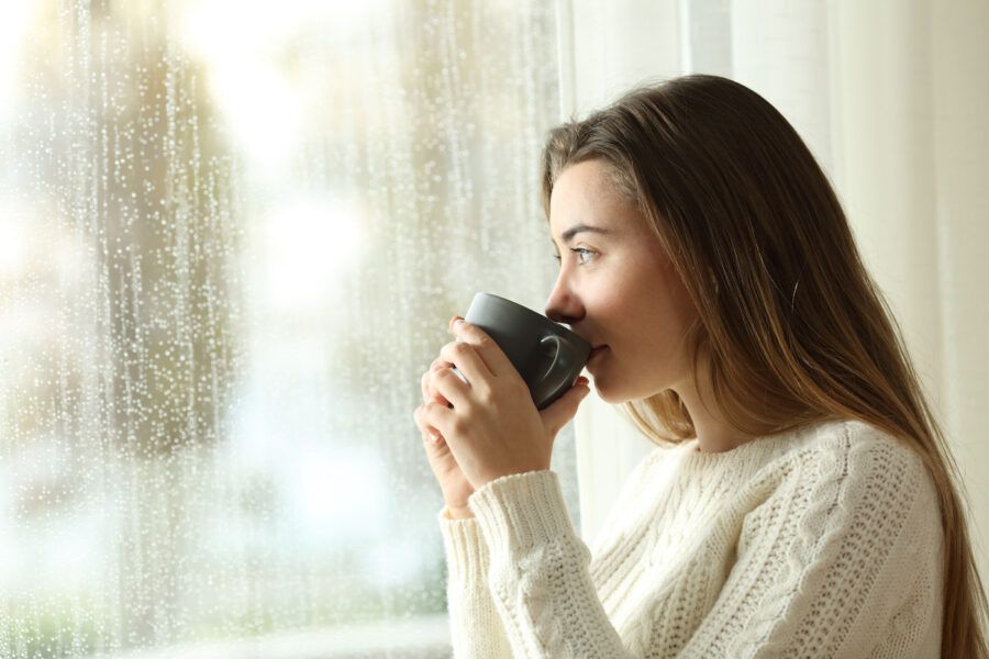 A woman drinking coffee looking through a window a rainy day