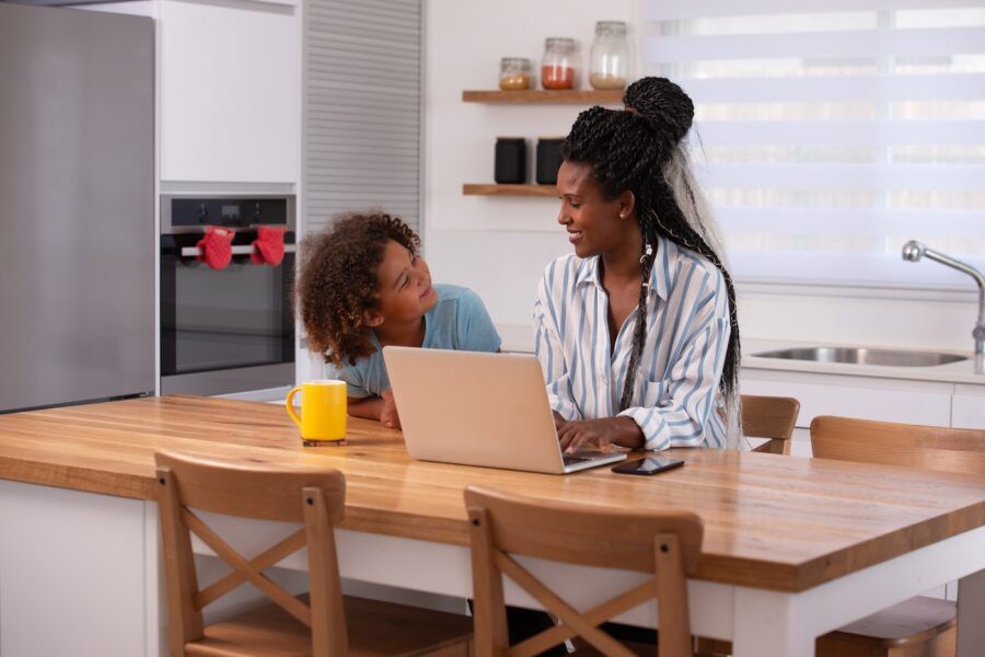 Freelancer woman working on laptop while sitting with daughter at the kitchen.