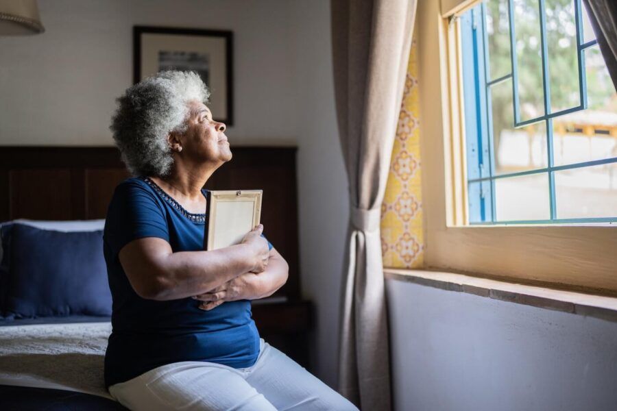 Older woman seated beside a bedroom window holding a framed photograph, with natural light streaming through the open window and a bed, curtains, and framed wall art visible in the background.