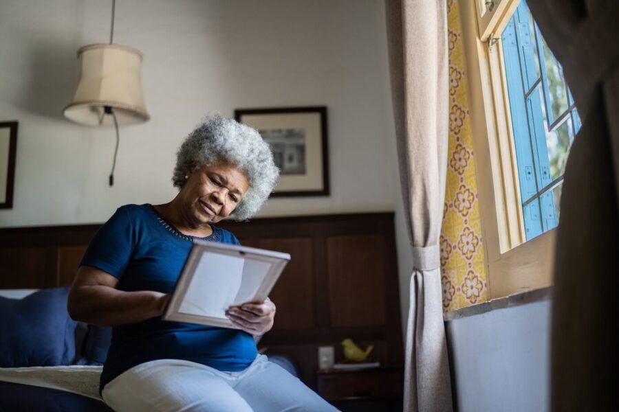 Older woman seated on the edge of a bed near a window, holding and looking at a framed photograph. The bedroom features wood-paneled furniture, framed artwork, patterned wall accents, and natural light streaming through an open window
