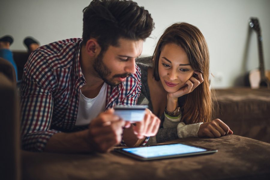 man and woman lying on brown couch looking at tablet screen. The man is holding a credit card.