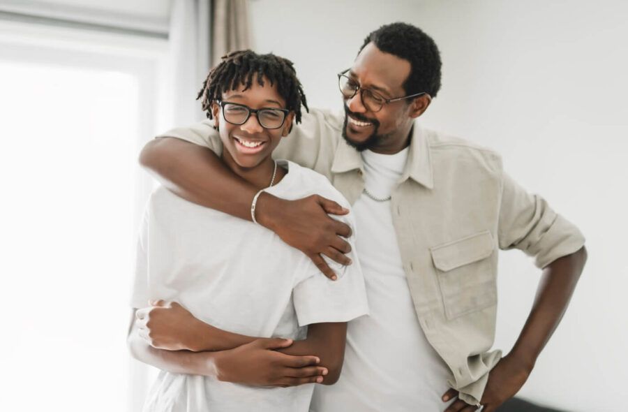 Adult with an arm around a teenager wearing glasses in a bright home setting, standing together near a window and looking toward the camera.