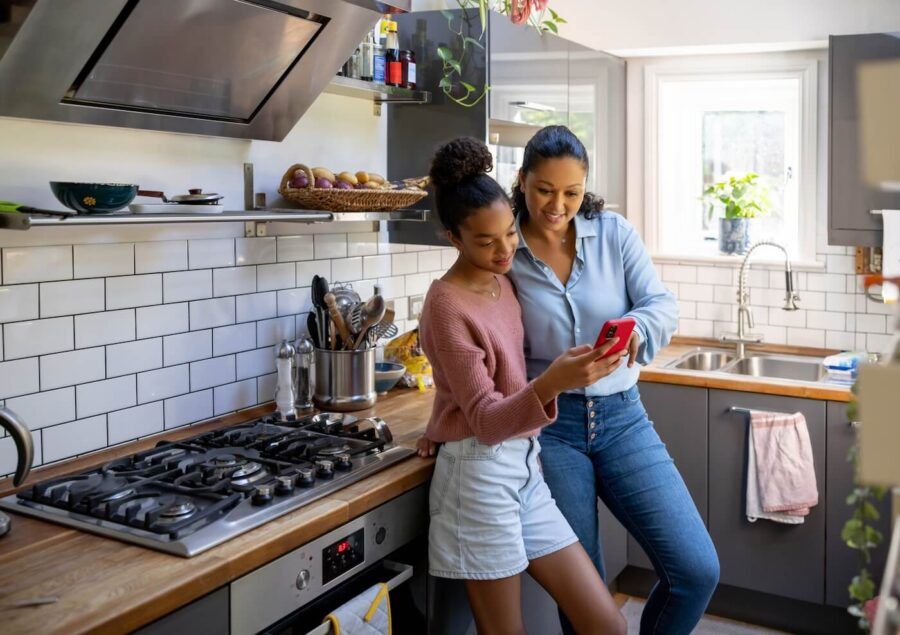 A woman and a teenage girl standing together in a bright kitchen, looking at a smartphone screen. The modern kitchen features wooden countertops, white subway tile backsplash, stainless steel appliances, and natural light from nearby windows.