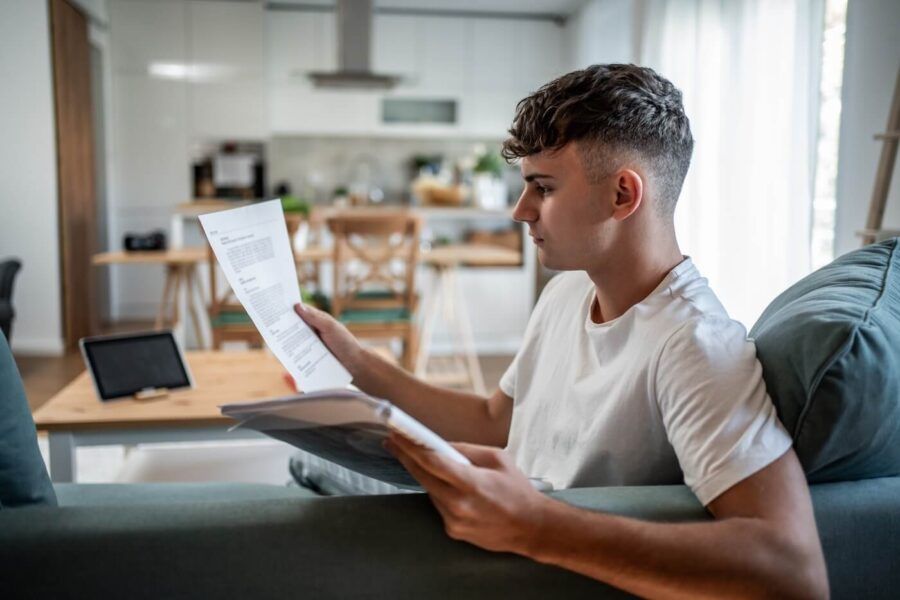 Focused young man sitting on a sofa and reviewing printed documents; a tablet and kitchen area are visible in the background.