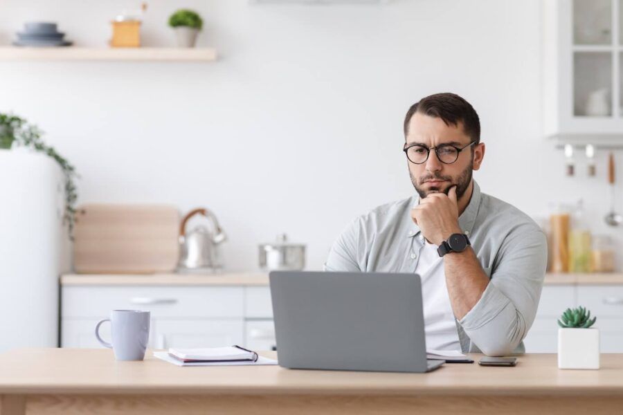 Focused man seated at a desk in a home office or kitchen workspace, looking at a laptop computer, with a notebook, smartphone, coffee mug, and small potted plant on the table.