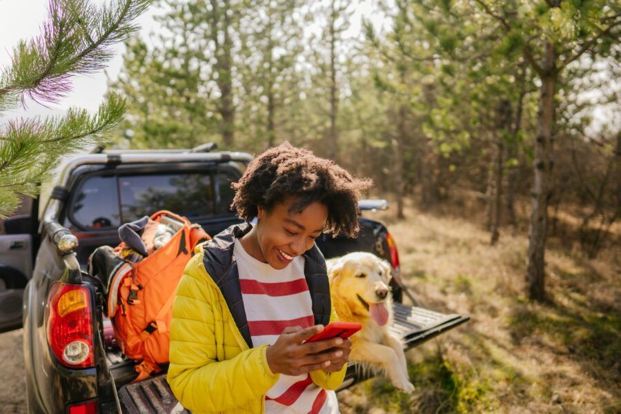 Photo of young smiling woman on a road trip with her retriever dog, riding along on their pick up truck; using her mobile phone to check auto insurance rates.