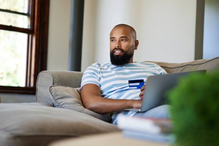 Relaxed man sitting on a sofa at home using a laptop while holding a credit card. Large windows, cushions, and home furnishings are visible in the background.