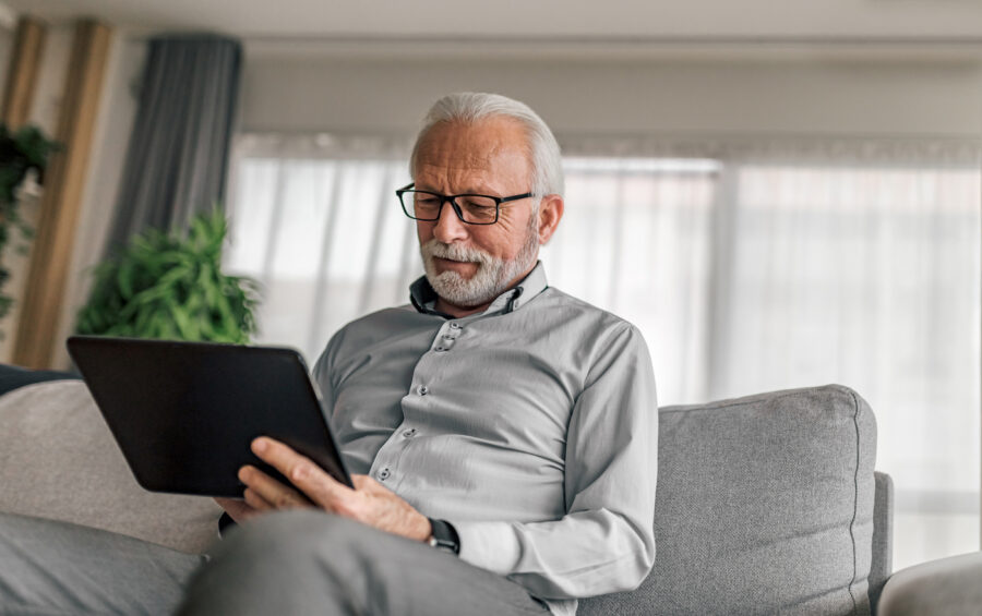 Elderly man using digital tablet while sitting on couch against window at home