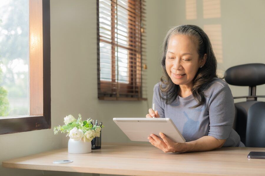 A woman in her 60s sitting by the window and using portable digital tablet to check retirement accounts.