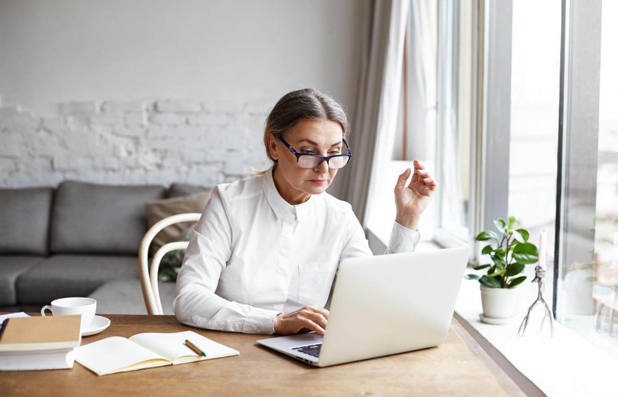 A senior woman wearing glasses is using her laptop at her desk next to a window in the living room.