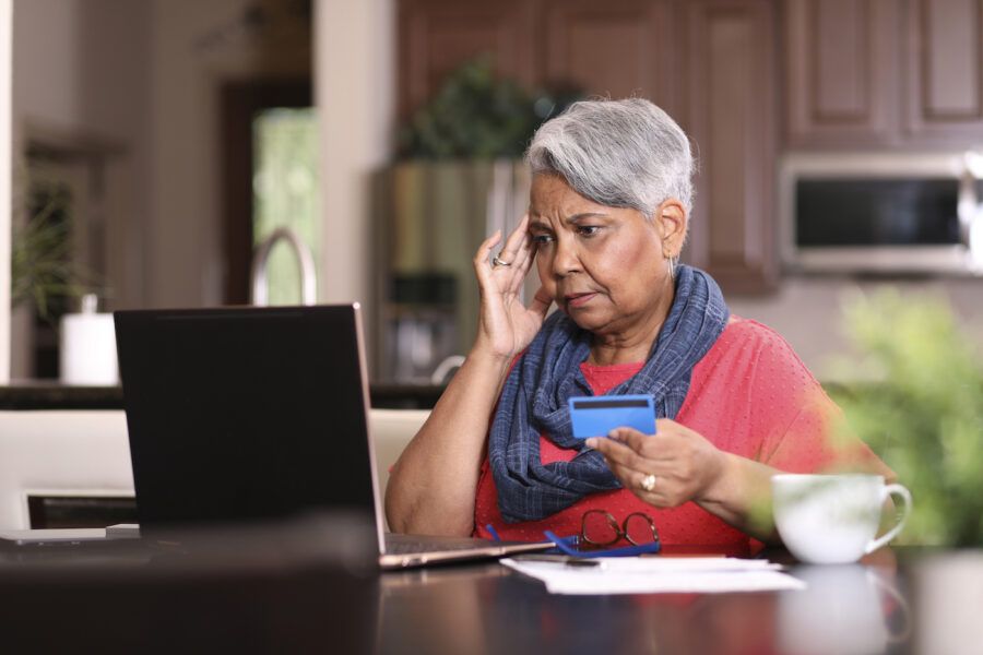 Senior woman looking at laptop and holding credit card is stressed about inflation.