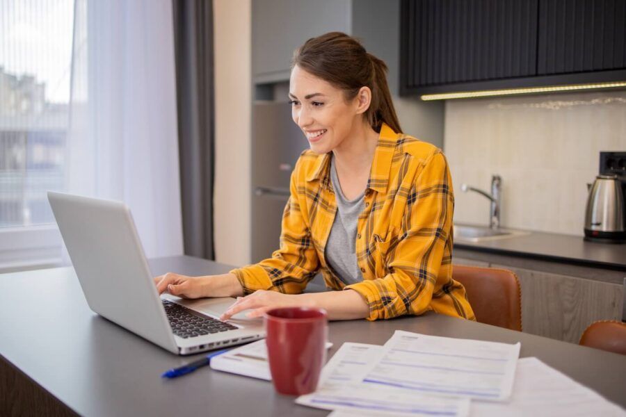 Smiling woman using her laptop in the kitchen with a red mug and printouts on the table