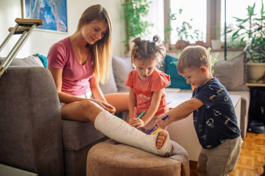 A mother with cast on her leg sitting on sofa, writing on the cast with her children, playful, enjoying and having fun