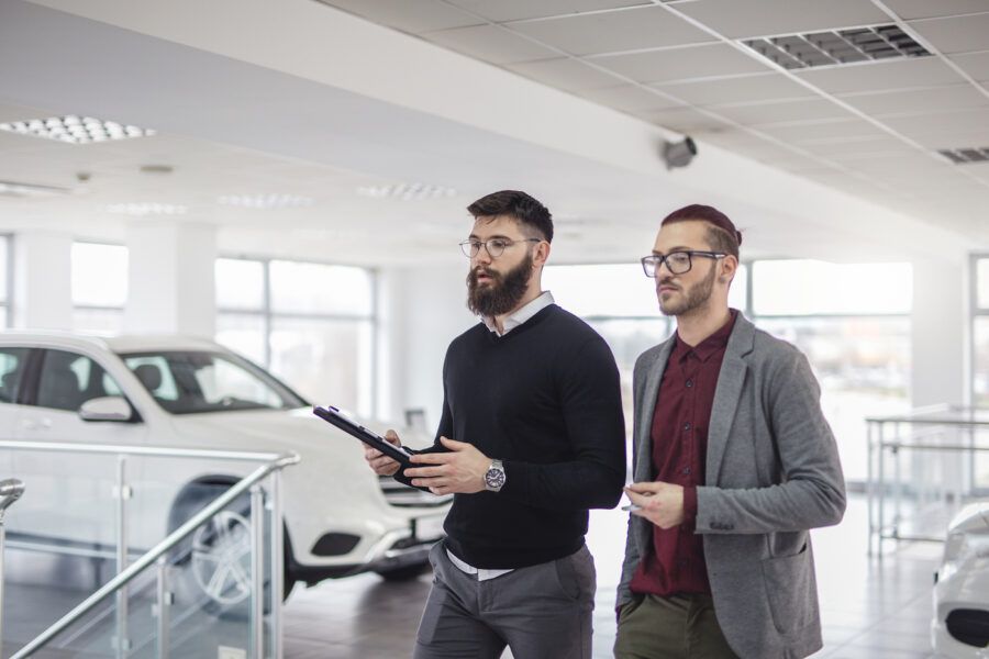 Young man with bad credit considering buying a new car from the car showroom, assisted by a salesman holding a clipboard.