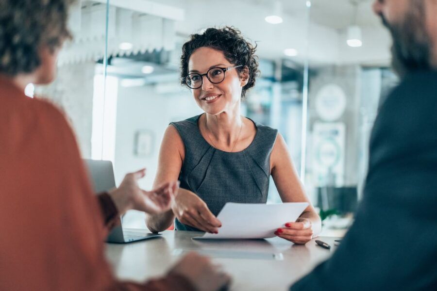 “Woman seated at a table in a modern office, holding papers and discussing documents with a couple during a meeting