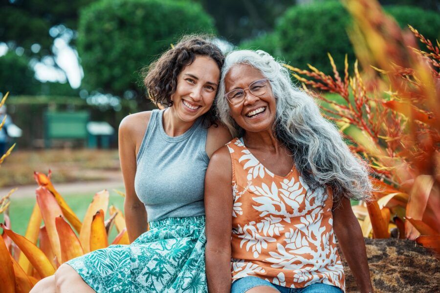 Portrait of a beautiful and healthy senior woman and her adult daughter smiling directly at the camera while sitting together outside surrounded by orange and yellow tropical foliage in Hawaii.