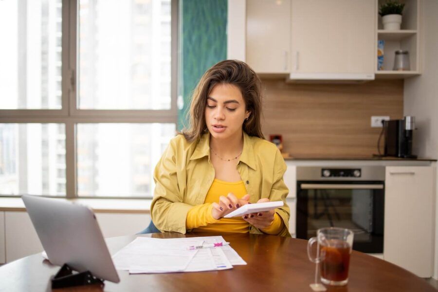 Focused young woman using a calculator while looking at the printouts on the table in front of her, with a tablet and a mug standing nearby