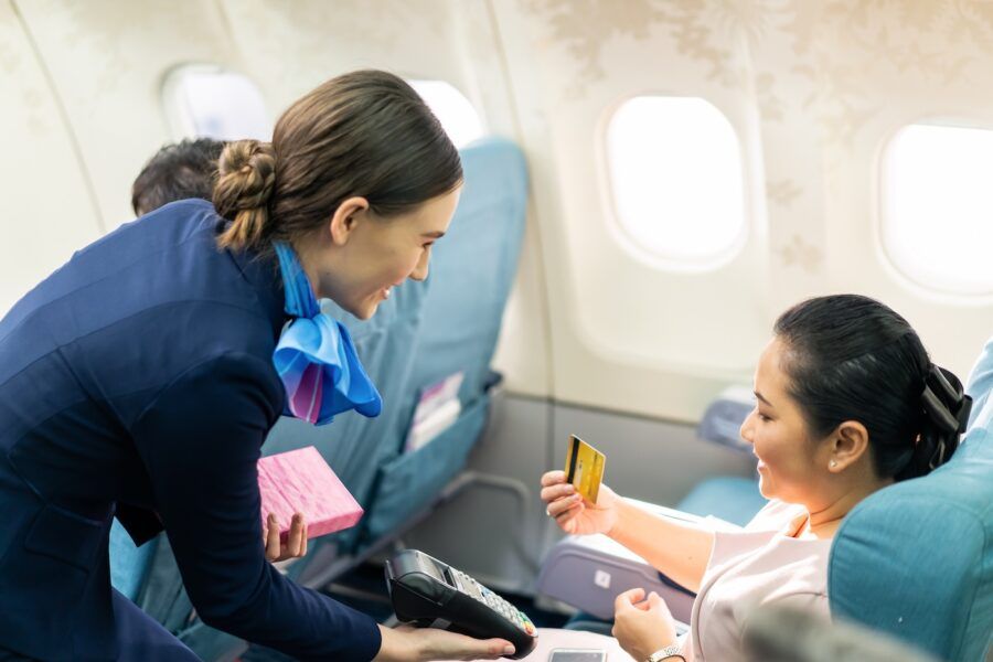 A woman holding a credit card seated on an airplane being helped by a flight attendant.