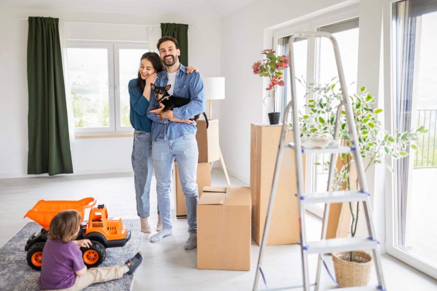 Young family stands inside a bright room with moving boxes, the man holding a small dog, while a child sits on the floor playing with a toy truck; a ladder and houseplants are visible nearby.